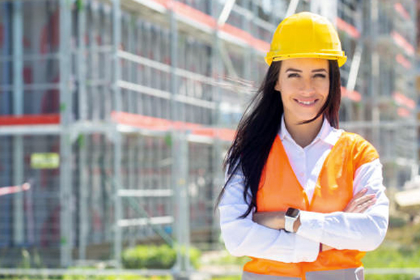 Female site supervisor wearing safety vest and helmet stands confidently in front of the construction covered in scaffolding.