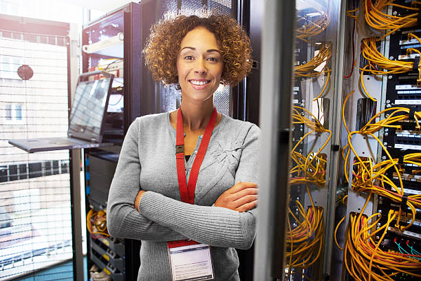 a female IT technician is editing the programme on the network server in a server room. She is looking to camera and smiling as she stands amongst the banks of servers and cables.