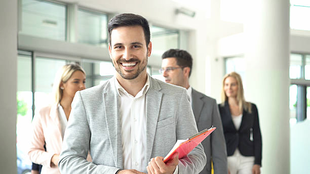 Closeup of handsome assertive businessman standing in front of his colleagues. He's smiling and looking at camera, wearing fray smart casual suit and holding a brochure.
