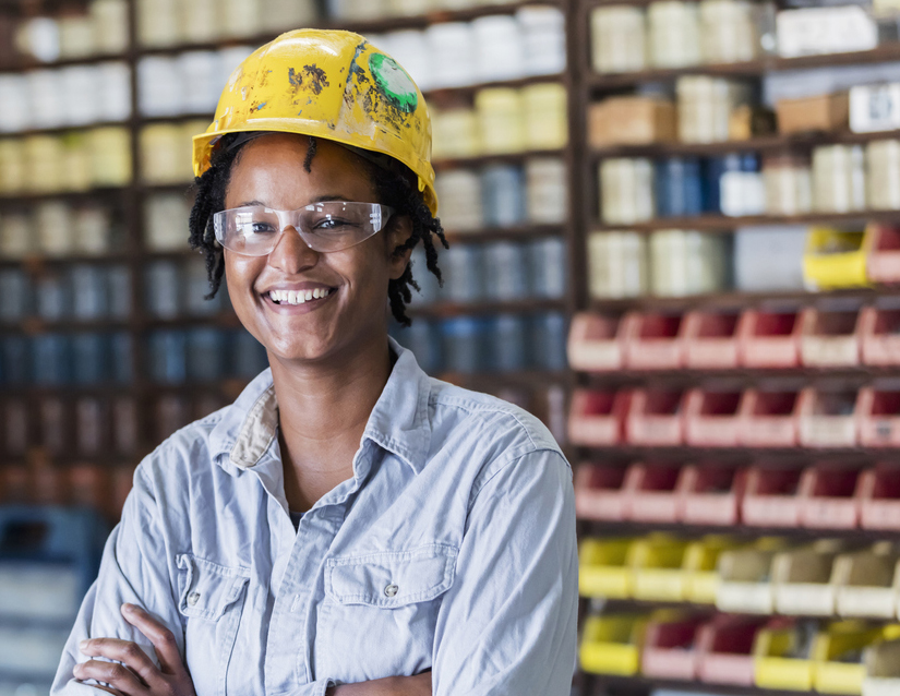 An black woman working in a printing plant, wearing a hard hat and safety goggles, smiling at the camera. She is standing in a storage warehouse, printing supplies in the background.