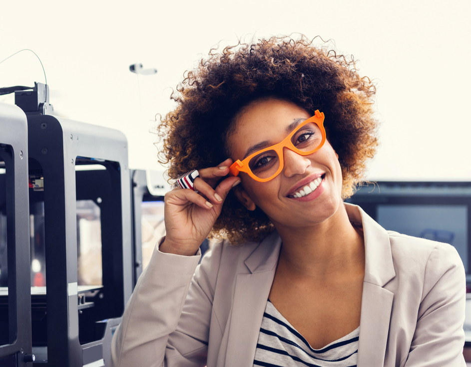 Mixed race friendly young woman wearing 3D printed glasses, sitting in a 3D printer office and smiling at the camera.