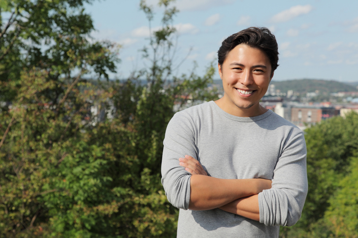 Attractive young man standing outside wearing a grey shirt. With a green city background.