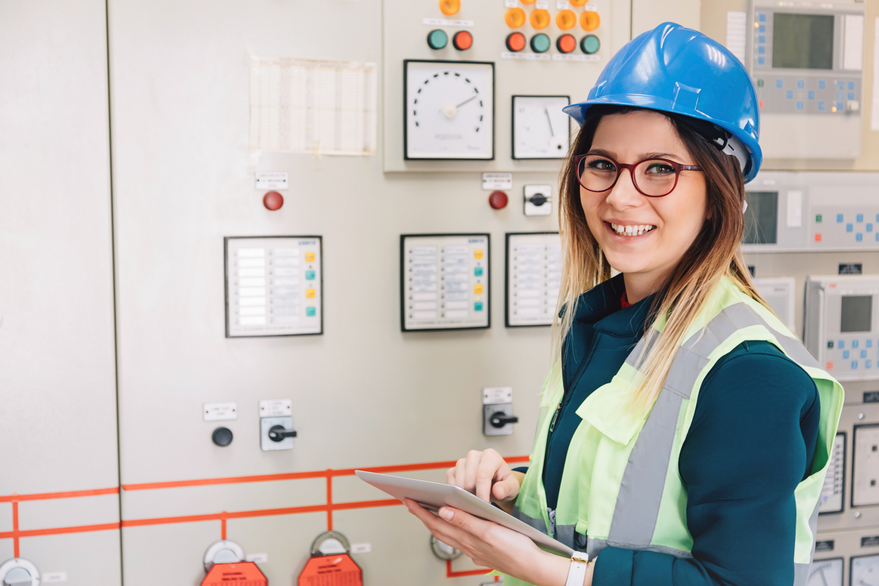 Portrait of young electrical engineer woman smiling and looking at camera posing while working on checking light indication status equipment at power plant with digital tablet pc in control room of modern electric energy station. XXXL size taken with Canon 5D MIV