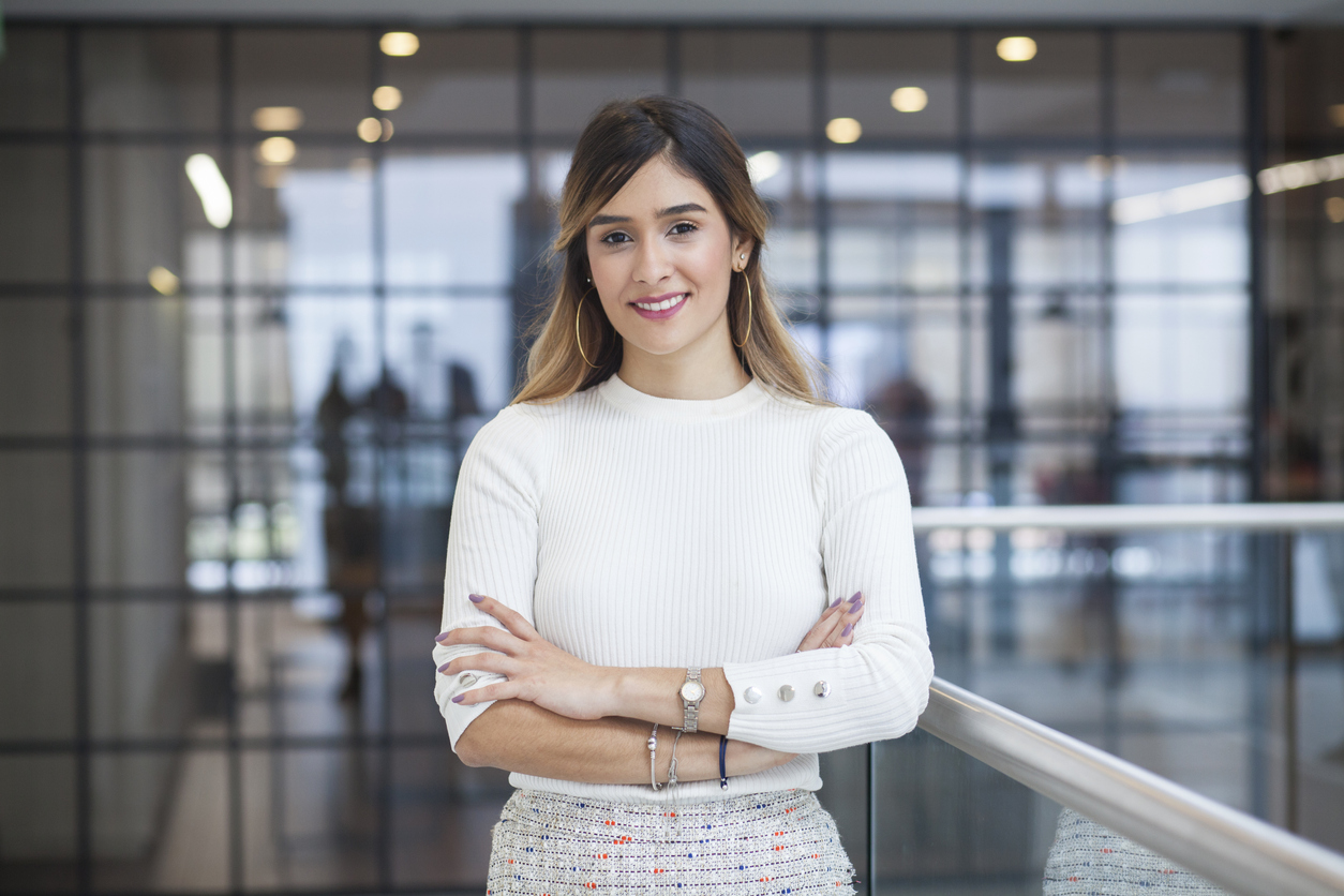 Executive Latin woman with long brown hair and white skirt and gray skirt with an approximate age of 28 is crossed arms and smiling at the camera with a half-sided stance in one of the corridors of the company for which she works next her a silver railing